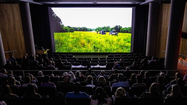 Theater full of people seen from the very back, looking toward a screen showing a grassy field.