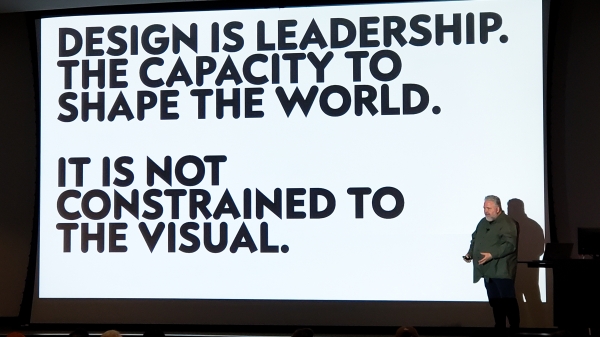 Designer Bruce Mau stands in front of a slide reading "Design is leadership. The capacity to change the world. It is not constrained to the visual."