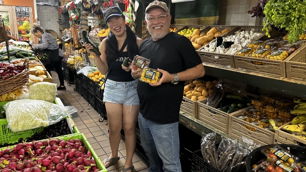 Two people among crates of fresh produce smile and hold up fruit and vegetables for the camera.