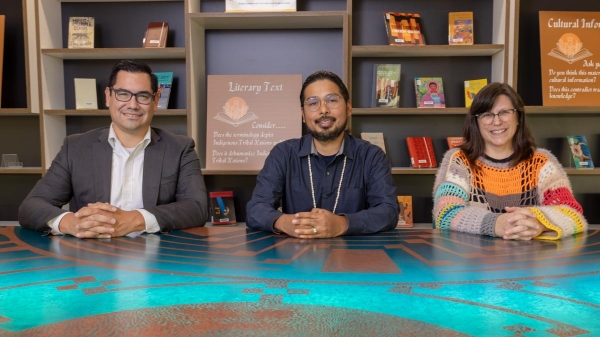 Alexander Soto, Trevor Reed and Lorrie McAllister sitting at the O'odham Storytelling table in Hayden Library, smiling for the camera with with bookshelves placed behind.